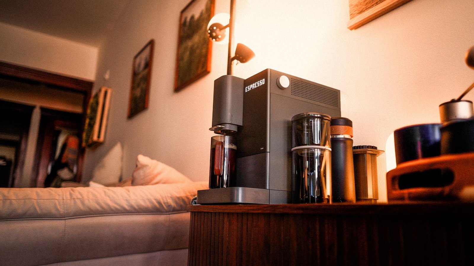 A dark gray Terra Kaffe Demi espresso machine on a walnut cabinet in a warm living room, with grinders and tumblers beside it