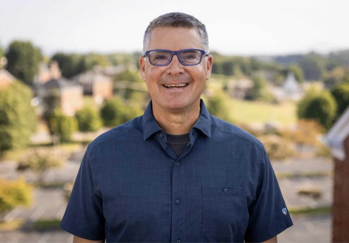 A smiling man in glasses and a navy shirt, outdoors above a small town